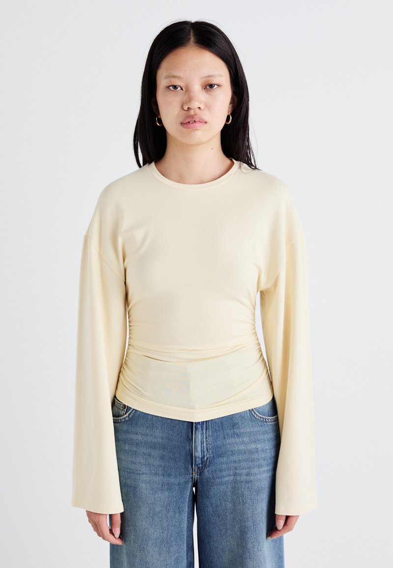Top lungo a maniche lunghe di colore giallo chiaro con scollo rotondo e dettagli arricciati in vita. Tessuto morbido e liscio. Indossato con jeans a gamba ampia blu.