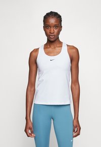 Bijela sportska majica bez rukava s širokim trakama i okruglim izrezom, s malim crnim Nike logotipom na sredini. Uparen s plavim tajicama.