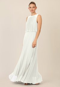 Mouwloze, vloerlange jurk van lichtwitte stof met geplooide textuur. Heeft een gelaagde, gedrapeerde bustier en een uitgeklapte rok.