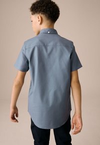 Lyhythihainen paita pehmeässä liuskekiven sinisessä kankaassa, jossa pyöristetty helma ja hillitty napinappi kauluksessa. Yhdistetty tummien housujen kanssa.