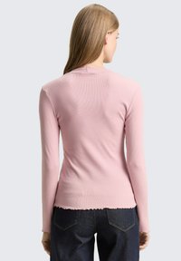 Long-sleeve pink ribbed top with a scalloped hem and fitted design, paired with dark denim jeans. The fabric has a smooth texture.