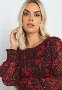 Svart blus med långa ärmar och röd blommönster, med texturerad tyg, smockade ärmslut och dekorativa silverdetaljer vid axlarna.