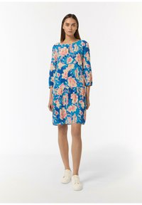 Blaues Blumenkleid mit pinken und beigen Blumen, Dreiviertelärmeln, einem runden Ausschnitt und einem gestuften Saum, kombiniert mit weißen Sneakers.