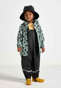 Vandtæt mintgrøn jakke med sorte prikker, sorte vandtætte bukser og gule gummistøvler. Inkluderer en sort bøttehat.