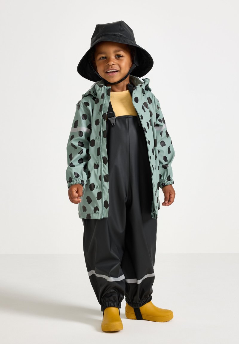Vandtæt mintgrøn jakke med sorte prikker, sorte vandtætte bukser og gule gummistøvler. Inkluderer en sort bøttehat.