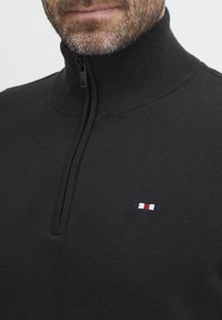 Suéter negro de punto con cuello alto y media cremallera. Presenta un pequeño logotipo bordado en rojo, blanco y azul en el lado izquierdo del pecho.