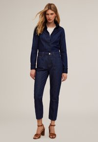 Mono vaquero en azul oscuro, con cuello tipo camisón, mangas largas y cintura ajustada. Destacado con cierres de botones y detalles en los puños.