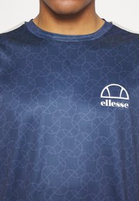 Granatowa koszulka z krótkim rękawem, z okrągłym wzorem, okrągłym dekoltem i białym logo 'ellesse' umiejscowionym na lewej piersi.