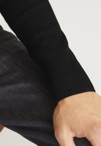 Svart ribbad tröjärm visad tillsammans med en hand, med en nära vy av den texturerade tyget och en mörk, slät bakgrund.