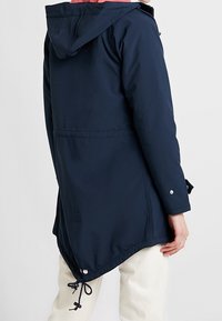 Marineblå vandtæt jakke med hætte, justerbar snor og sidelommer; har en længere rygstykke og en glat tekstur.