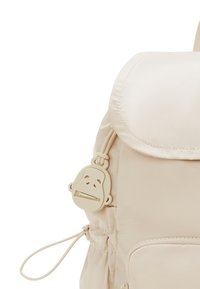 Beige Rucksack mit Klappdeckel, vorderer Reißverschlusstasche und einem affenförmigen Reißverschlussanhänger an der Seite.
