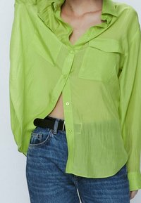Lime green sheer button-up shirt with two front pockets, relaxed fit, and rolled sleeves, paired with dark blue denim jeans and black belt.