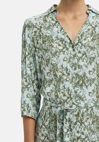 Robe à motifs floraux en bleu clair et vert olive, fabriquée en tissu doux. Comprend un col, une fermeture à boutons et une ceinture à la taille. Manches trois quarts.