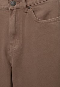 Gros plan d'un pantalon en denim marron avec un bouton en métal, une poche avant et des détails de couture visibles sur le tissu.