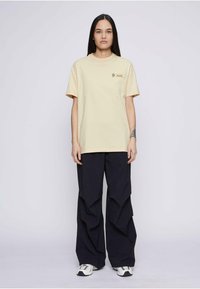 Camiseta de algodón beige con mangas cortas y un logo en el pecho, combinada con pantalones de pierna ancha azul oscuro con diseño en capas y zapatillas blancas.