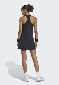 Frau in schwarzem ärmellosem Tennis-Kleid, weißen Socken, grau-weißen Turnschuhen und schwarzen Arm- Bands, die mit dem Rücken zum Betrachter steht.