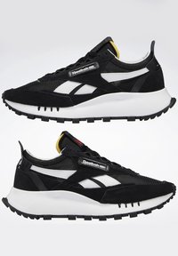 Baskets Reebok noires et blanches avec une tige en mesh et en suède, une semelle blanche coussinée, et une étiquette avec le logo Reebok sur le côté.