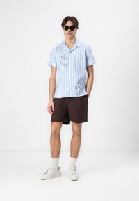 Helles blaues Kurzarmhemd mit vertikalen Streifen und einem Oktopus-Print, kombiniert mit braunen Shorts und weißen Sneakers, vor einem neutralen Hintergrund.