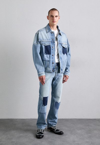 Man standing against plain wall wearing light blue denim jacket and jeans with dark patches and heart-shaped designs, black dress shoes.