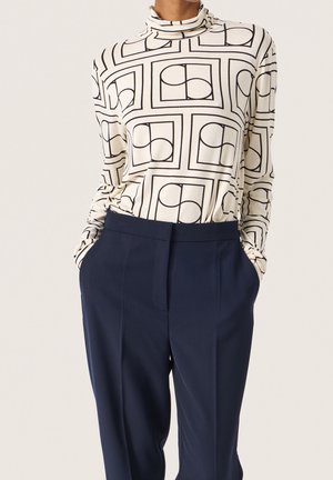Pitkähihainen kerma-toppi mustalla geometrisella kuviolla. Yhdistetty tummansinisiiin korkeavyötäröisiin housuihin, joissa on sivutaskut ja räätälöity istuvuus.