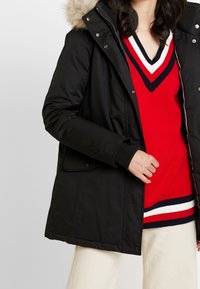 Parka nera isolata con cappuccio in pelliccia removibile, caratterizzata da una chiusura con zip, tasche laterali e polsini a costine, indossata sopra un maglione a righe rosse.
