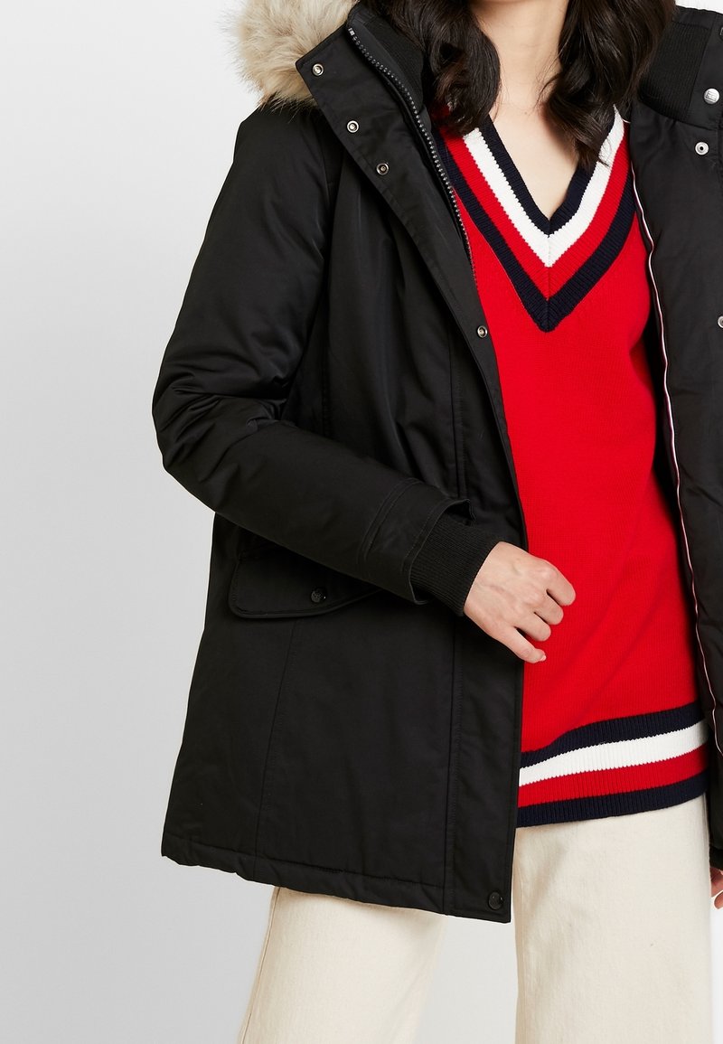 Parka nera isolata con cappuccio in pelliccia removibile, caratterizzata da una chiusura con zip, tasche laterali e polsini a costine, indossata sopra un maglione a righe rosse.