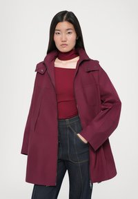 Veste bordeaux avec un col haut, fermeture éclair à l'avant et poches latérales. Assortie à un haut coordonné et un pantalon en denim foncé. Texture lisse.