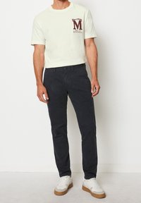 Outfit maschile con una T-shirt beige chiaro con un logo marrone scuro e chinos blu scuro, abbinata a sneakers bianche con dettagli beige.