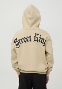 Felpa con cappuccio beige con la scritta "Street King" in bold nero sulla schiena. Materiale in cotone, vestibilità comoda e polsini a costine.