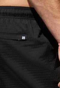 Fekete sportnadrágok texturált felülettel, rugalmas derékkal és egy apró cipzáras zsebbel az oldalon. Diszkrét logó részlet található rajtuk.