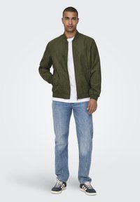 Bomber verde oliva in morbido materiale scamosciato con cerniera frontale, polsini e orlo a coste, indossato sopra una camicia bianca e jeans blu.