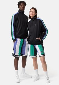 Jachetă neagră cu fermoar, cu accente în dungi albastre și verzi; pantaloni scurți asortați, cu dungi în nuanțe de teal, violet și negru. Adidași albi.