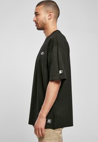 Black cotton t-shirt with short sleeves, loose fit, featuring white logo accents on the chest and sleeve, textured fabric, and side label.