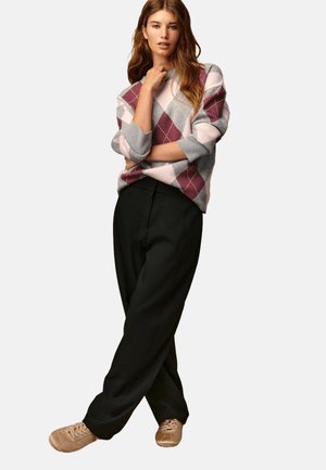 Jonge vrouw met lang haar in een roze en grijs ruitjestrui, zwarte wijde broek en beige sneakers, staand met gekruiste benen.