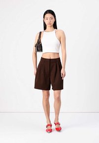 Hvit crop top, brune skreddersydde shorts og røde spisse sko. Modellen holder en liten svart håndveske med merkbare detaljer i maskinvare.