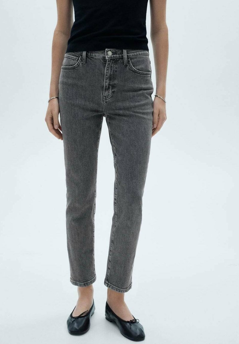 Personne portant un jean gris taille haute à jambes droites, un haut noir sans manches, des bracelets argentés et des ballerines noires sur un fond clair uni.