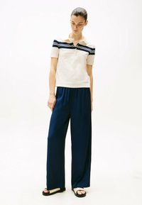 Beige polo-shirt med navy vandrette striber, korte ærmer og tre knapper. Løs pasform navy bukser lavet af glat stof og sorte sandaler.