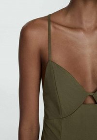 Žena v olivově zelených šatech bez rukávů s tenkými ramínky a malým výřezem pod poprsím, který odhaluje její ramena a horní část hrudníku.