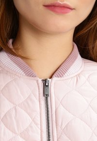 Nahaufnahme einer Person, die eine hellrosa gesteppte Jacke mit einem silbernen Reißverschluss und einem gerippten Kragen trägt. Es wird das untere Gesicht und der Hals gezeigt.