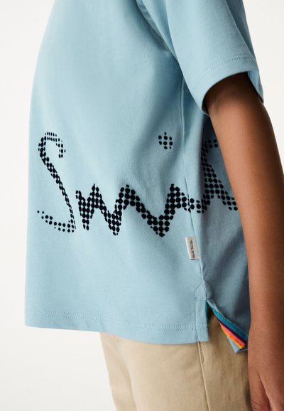 Gaiši zils kokvilnas T-krekls ar īsām piedurknēm, kas rotāts ar melnu punktiņu grafiku un sānu šķēlumu, caur kuru redzams daudzkrāsains apakšmalas galiņš.