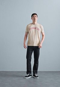Beige T-Shirt mit rotem "DSQUARED2"-Text, dunkle Jeans und schwarze Sneaker. Das Shirt weist einige Flecken auf und vermittelt einen lässigen Look.
