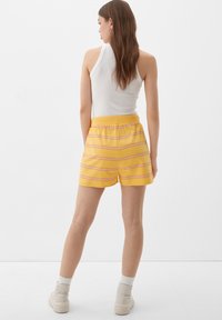 Gelbe Shorts mit rosa Streifen, elastischem Bund und lässiger Passform. Kombiniert mit einem weißen gerippten Tanktop und knöchellangen beigen Stiefeln.