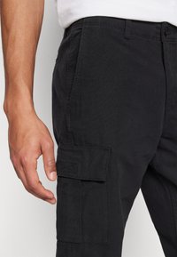 Pantalones cargo negros de algodón, con un bolsillo lateral, corte recto y detalles de costura mínimos. Primer plano de la textura de la tela.
