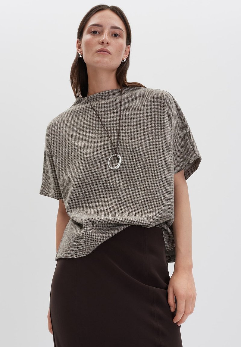 Frau in einem lockeren grauen strukturierten T-Shirt mit kurzen Ärmeln, schwarzem Rock, silbernen Creolen und einer langen Anhängerkette vor einem schlichten Hintergrund.