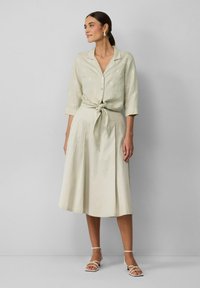 Lichtgroen linnen overhemd met bloemendruk, vastgeknoopt in de taille, gecombineerd met een vloeiende crèmekleurige rok. Model draagt witte sandalen met bandjes.
