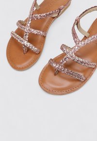 Sandalias planas con tiras trenzadas metálicas rosas, una suela de cuero marrón y un diseño minimalista. Presentan detalles de tiras cruzadas para un ajuste seguro.