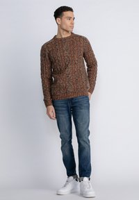 Brown knit sweater with textured vertical stripes, round neckline, paired with blue jeans and white sneakers. Model stands against a light background.