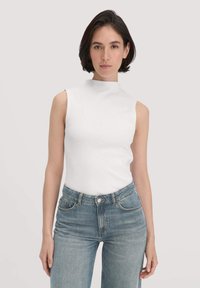 Vrouw met kort donker haar die een mouwloos wit coltrui-topje en een blauwe spijkerbroek draagt, staand tegen een effen lichte achtergrond.