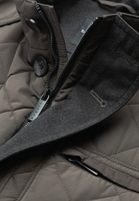 Quilted brown jacket featuring a black collar, zip closure, button accents, and a fabric tag. Textured materials and contrasting colors.