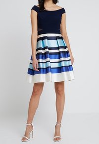 Bodice bez ramínek v námořnické modré, rozšířená sukně s modro-bílými vodorovnými pruhy. Hladká látka, délka nad kolena, doplněno bílými podpatky.